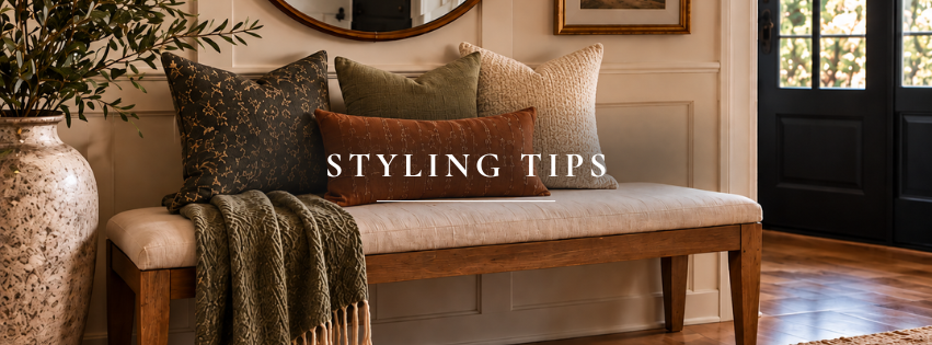 Warm entryway with wood bench, layered pillows, olive throw, round mirror, and black front door